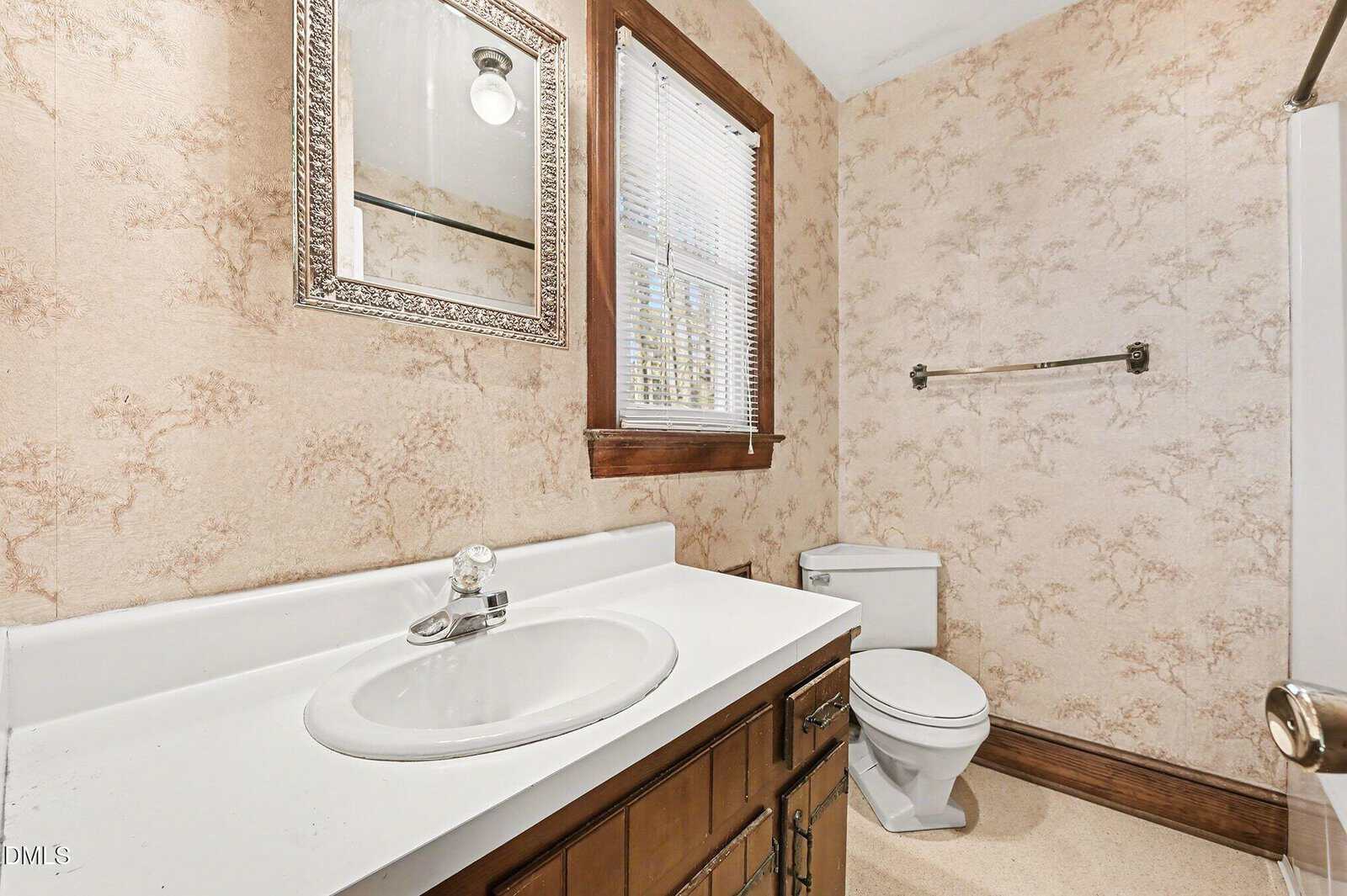 A bathroom with beige patterned wallpaper, a white sink with brown vanity, a wall-mounted mirror, a window with blinds, a toilet, a towel bar, and part of a shower or tub visible on the right.