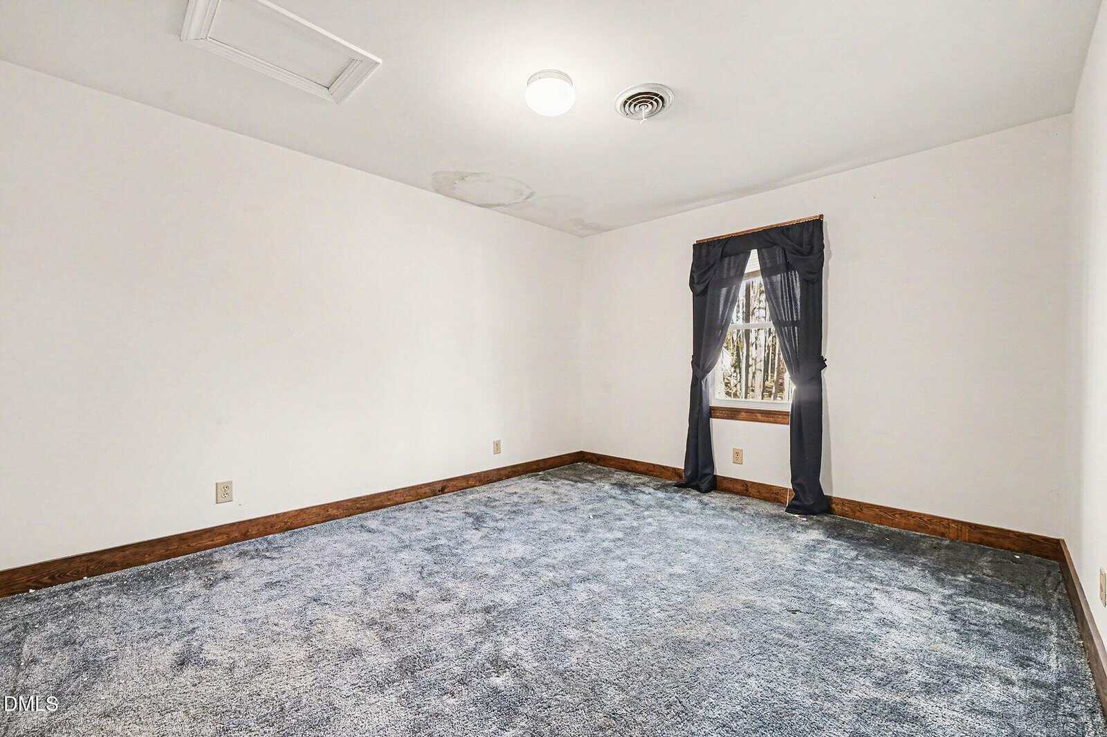 Empty room with white walls, blue carpet, a single window with dark curtains, wood trim, and visible water damage on the ceiling. Natural light enters through the window.