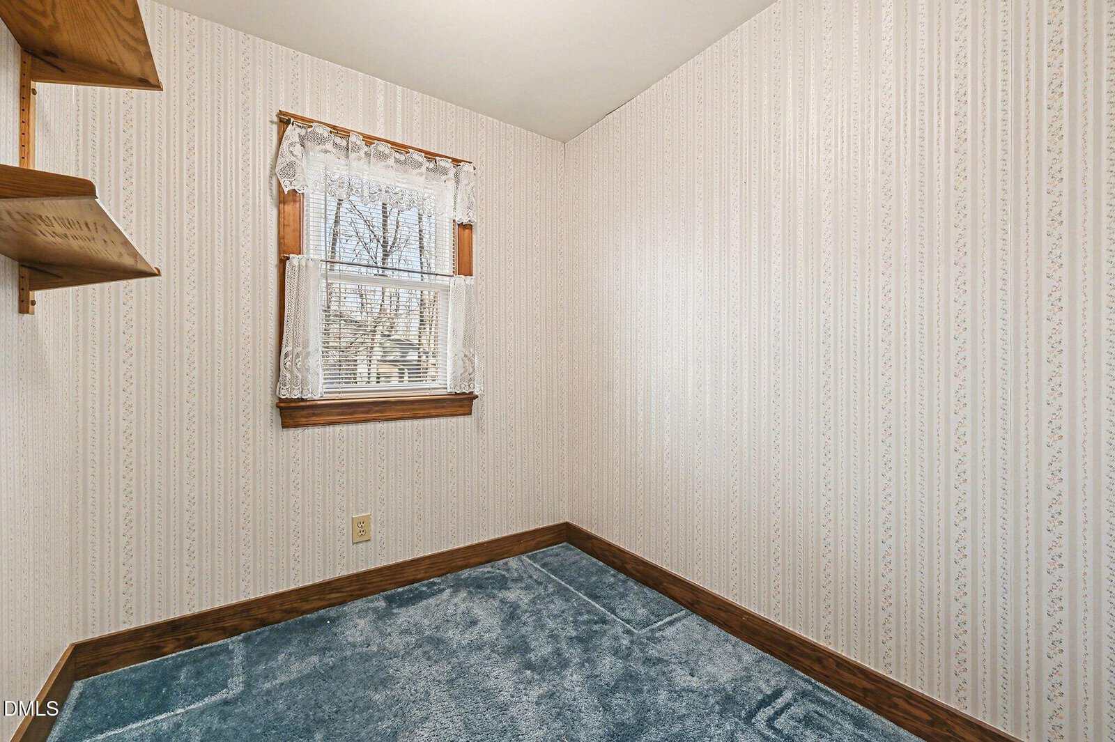 Small room with blue carpet, light wallpaper, wooden shelves on the left wall, and a window with lace curtains letting in natural light.