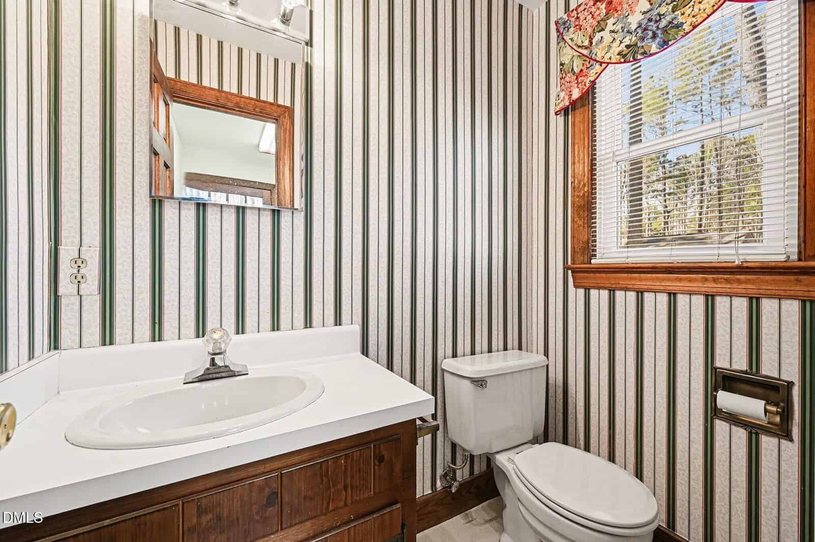 A small bathroom with vertical striped wallpaper, a white sink with a wooden vanity, a wall-mounted mirror, a toilet, a window with blinds and a floral valance, and a toilet paper holder.