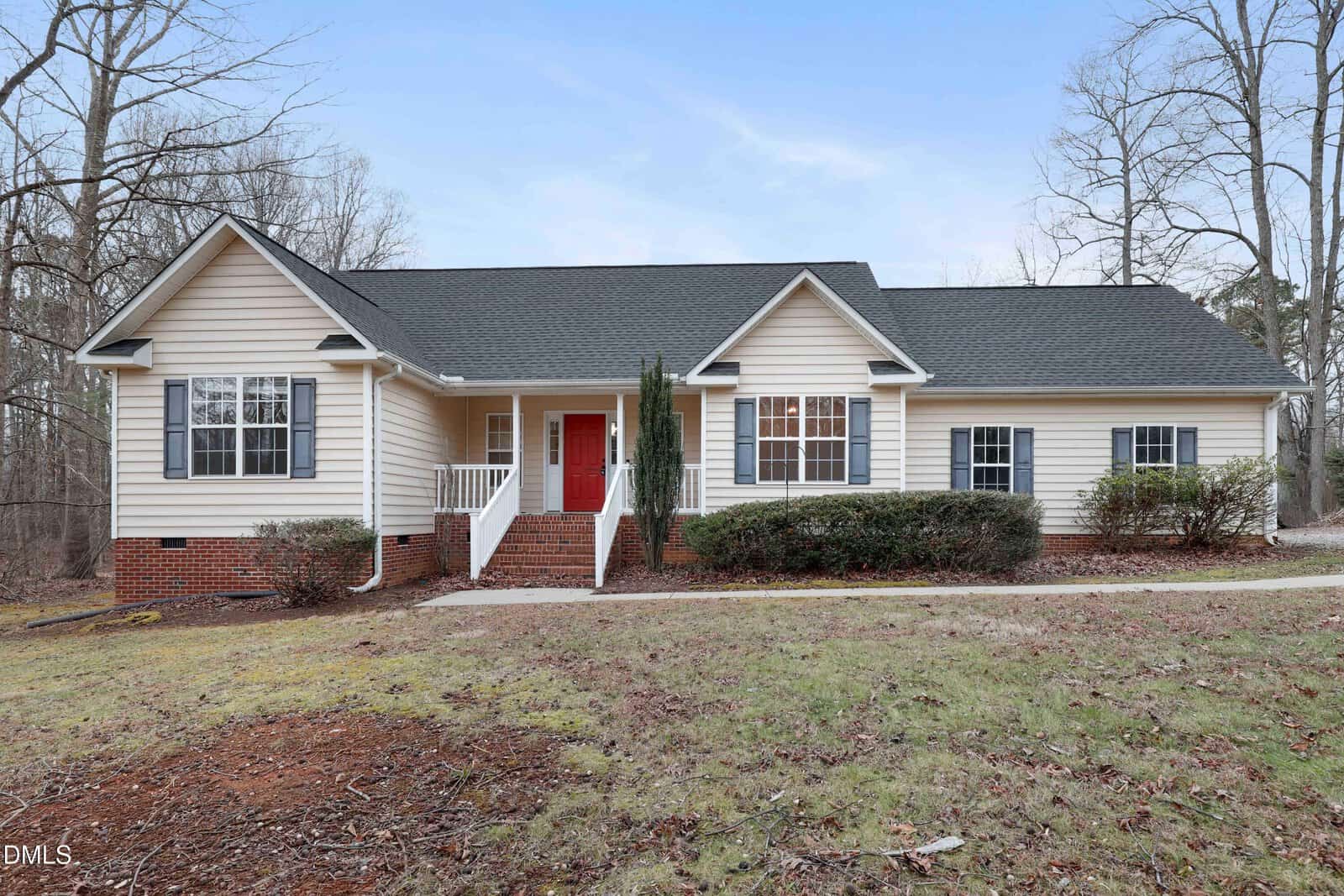 Charming single-story house in Oxford with light-colored siding, a dark shingled roof, red front door, brick foundation, white railings, and front steps. The yard at 2188 Elliott Street features bare soil, sparse grass, and trees in the background.