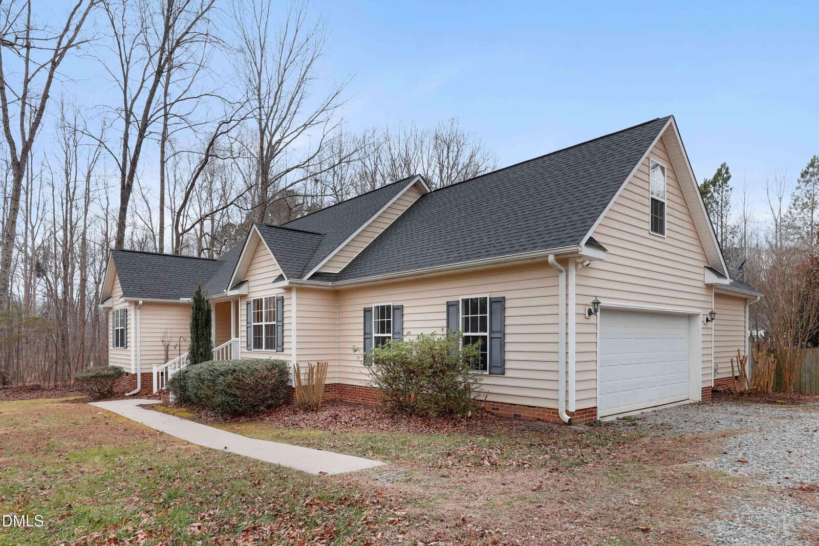 Single-story beige house with black shutters, a dark roof, and attached two-car garage. The house is surrounded by leafless trees and has a concrete walkway leading to the front porch.