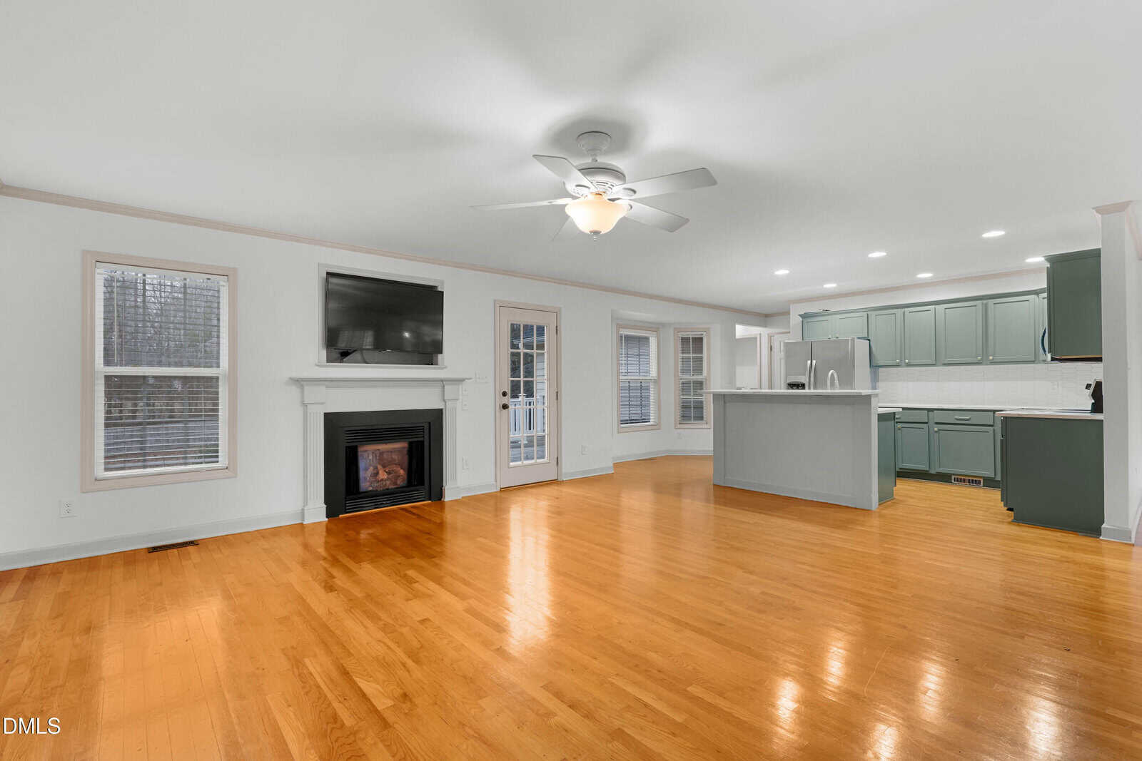 Open-concept living area with hardwood floors, a ceiling fan, fireplace with TV above, and adjoining kitchen featuring green cabinets, stainless steel appliances, and recessed lighting. Multiple windows let in natural light.