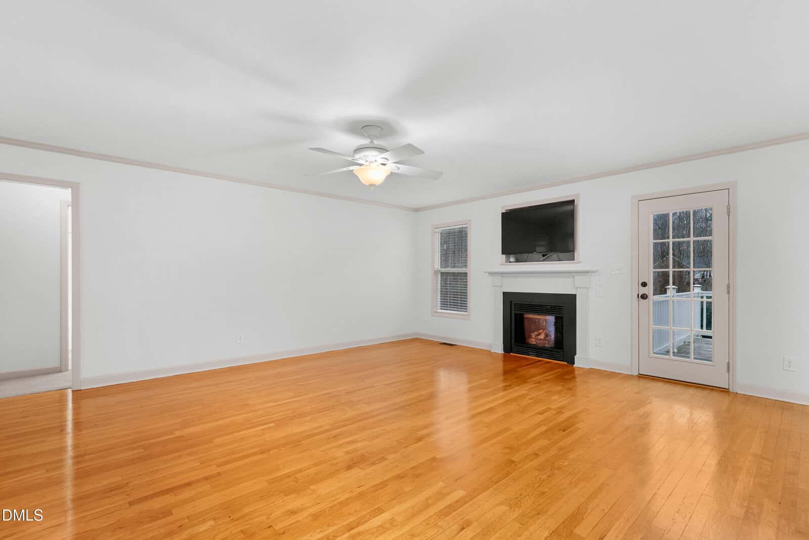 A spacious living room with light hardwood floors, white walls, a ceiling fan, a fireplace with a TV above it, two windows, a glass door, and visible entryways to other rooms.