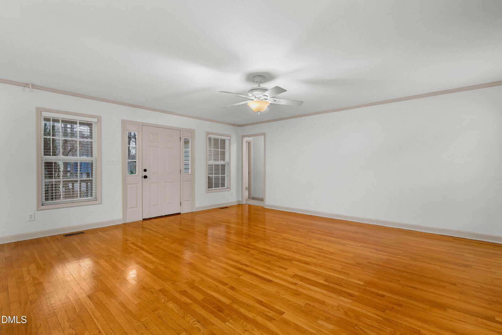 Empty living room with polished hardwood floors, white walls, two windows, a white front door with glass panels, ceiling fan with light, and an open doorway leading to another room.