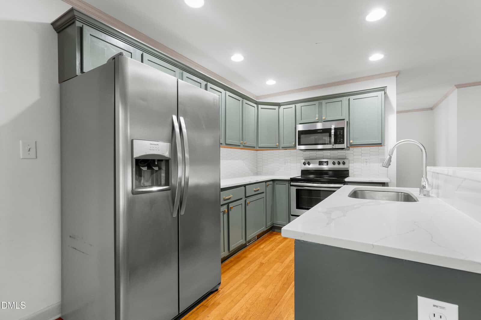 A modern kitchen with stainless steel appliances, green cabinets, white countertops, and wood flooring. The refrigerator, oven, microwave, and sink are visible, with recessed ceiling lights providing illumination.
