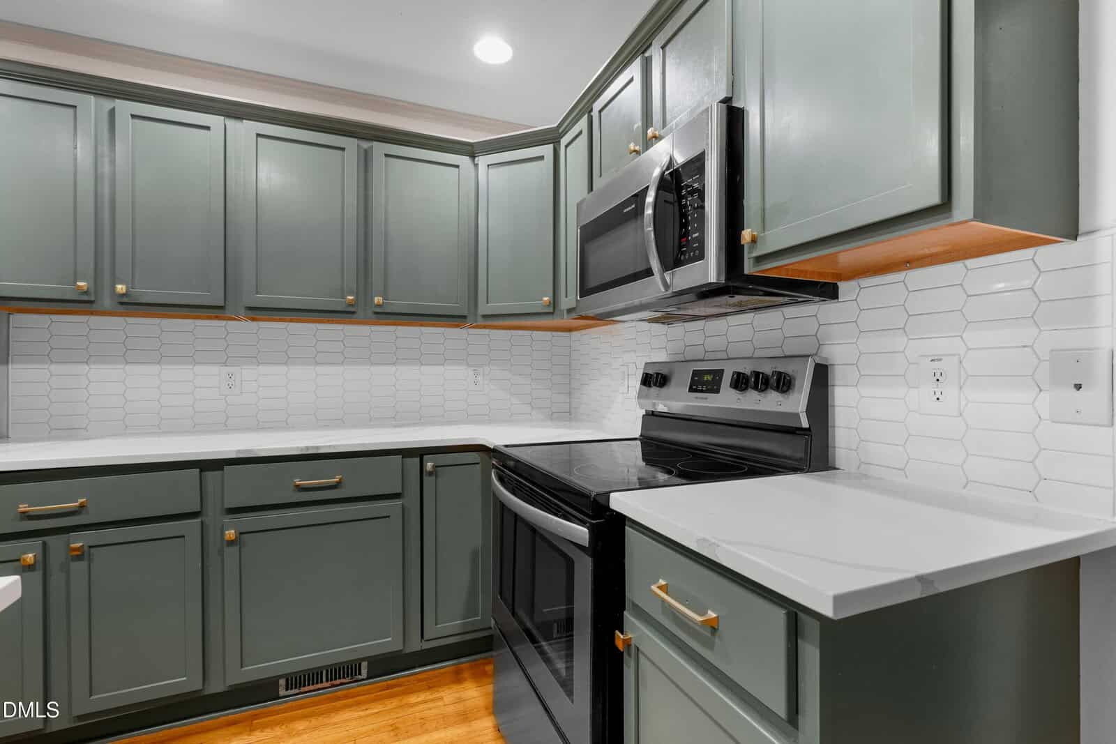A modern kitchen with green cabinets, white hexagonal tile backsplash, stainless steel appliances, white countertops, wood flooring, and brass hardware on the cabinets.