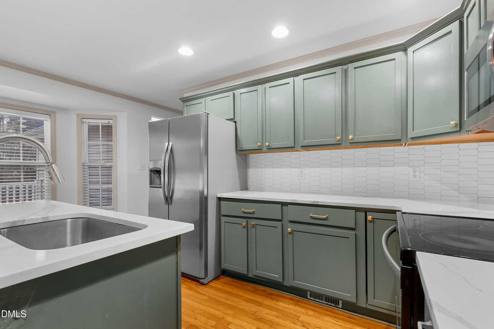 A kitchen with light green cabinets, white countertops, a stainless steel refrigerator, an electric stove, a sink with a curved faucet, white tile backsplash, and wood flooring. Recessed lights are on the ceiling.