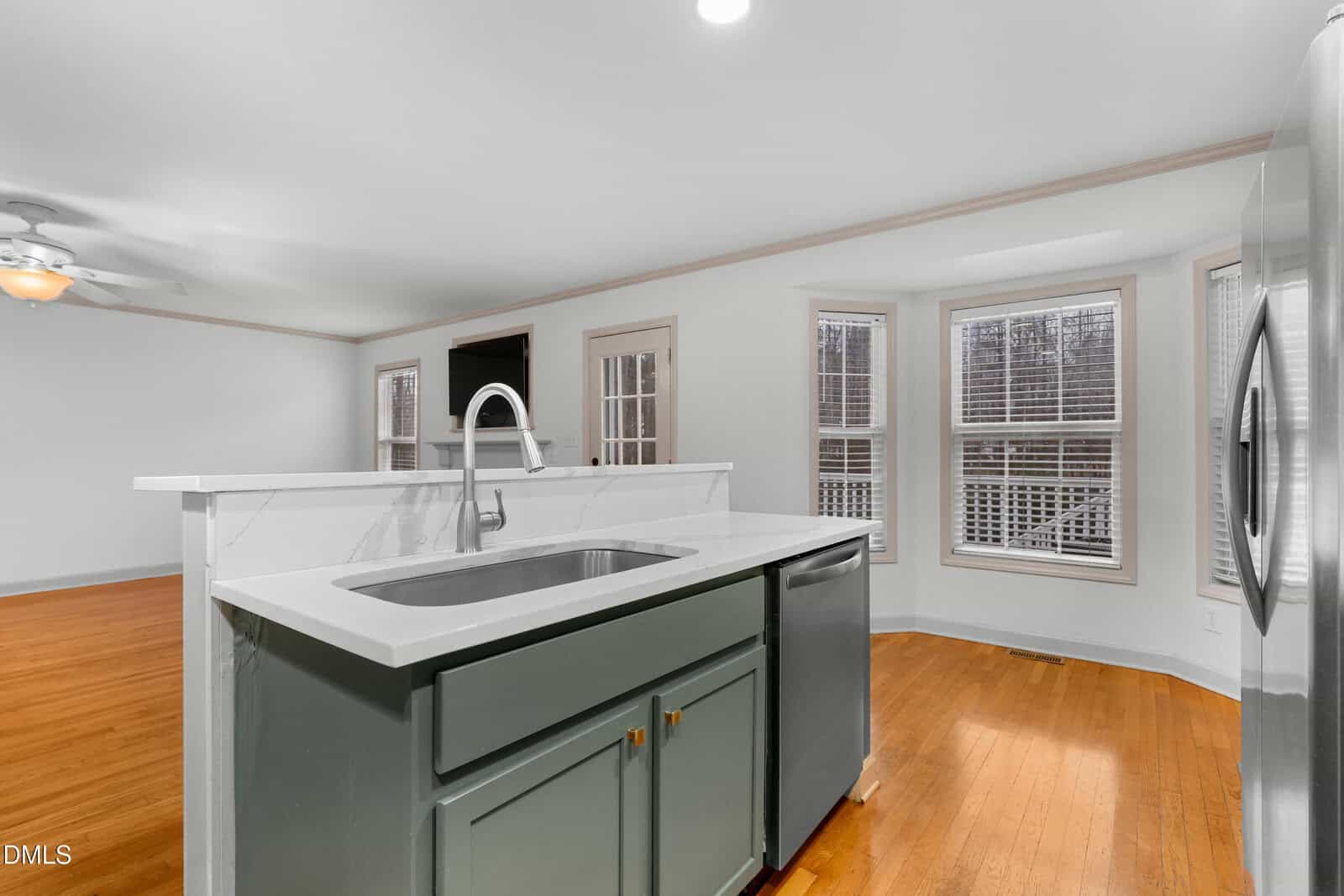 A modern kitchen with a green island featuring a sink and dishwasher, stainless steel refrigerator, hardwood floors, and a bright dining area with bay windows and white blinds.