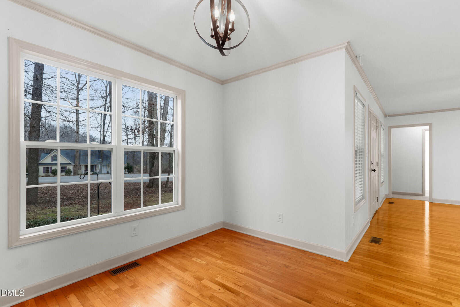 A bright, empty room with white walls, hardwood floors, a large window overlooking trees and a house, a modern chandelier, and a hallway leading to other rooms.