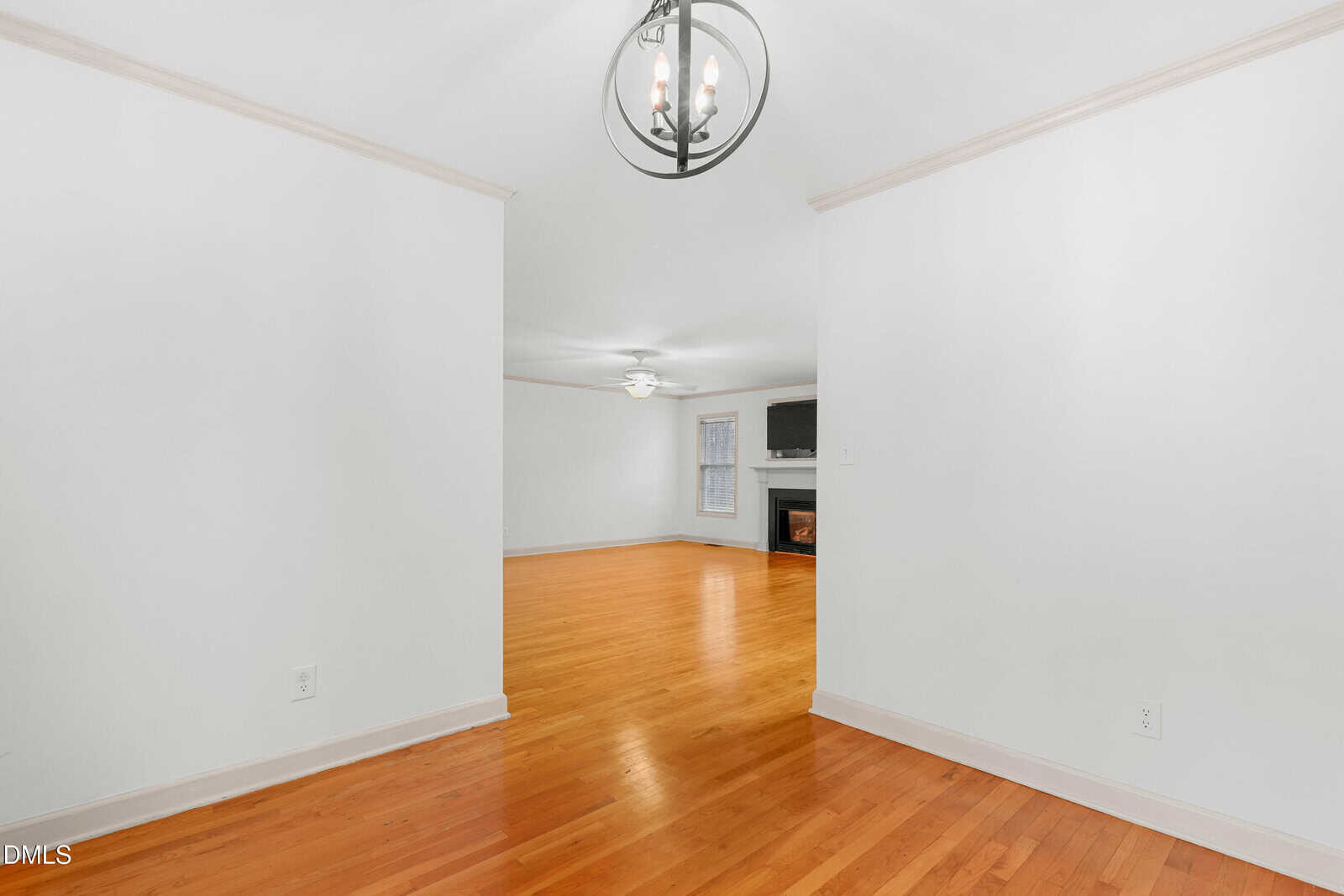 A bright, empty room with white walls and light hardwood floors. A chandelier hangs from the ceiling, and an open doorway leads to a living area with a fireplace and a ceiling fan.