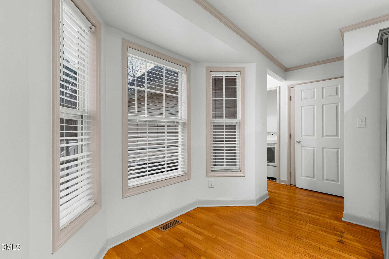 A bright room with three large windows, white walls, hardwood floors, and white blinds; a doorway shows a glimpse of a laundry area with a washer and dryer.