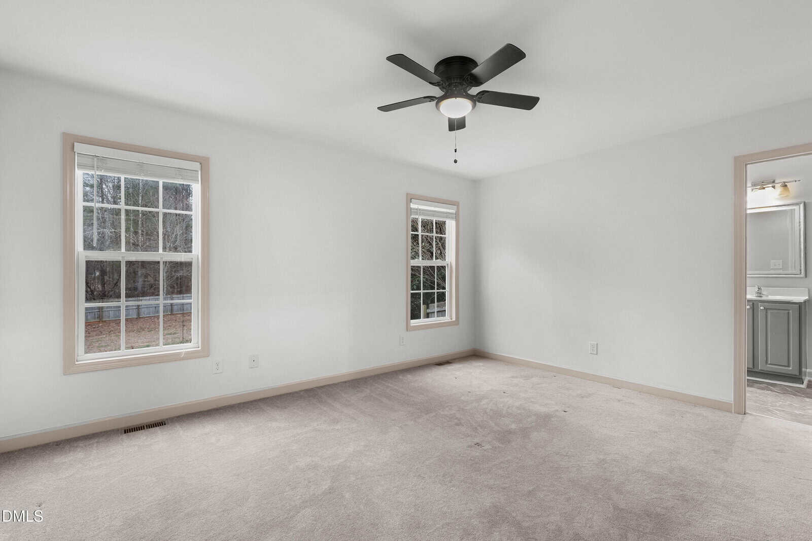 Empty carpeted room with two windows, white walls, beige trim, a black ceiling fan with a light, and a view into a bathroom with a gray vanity.
