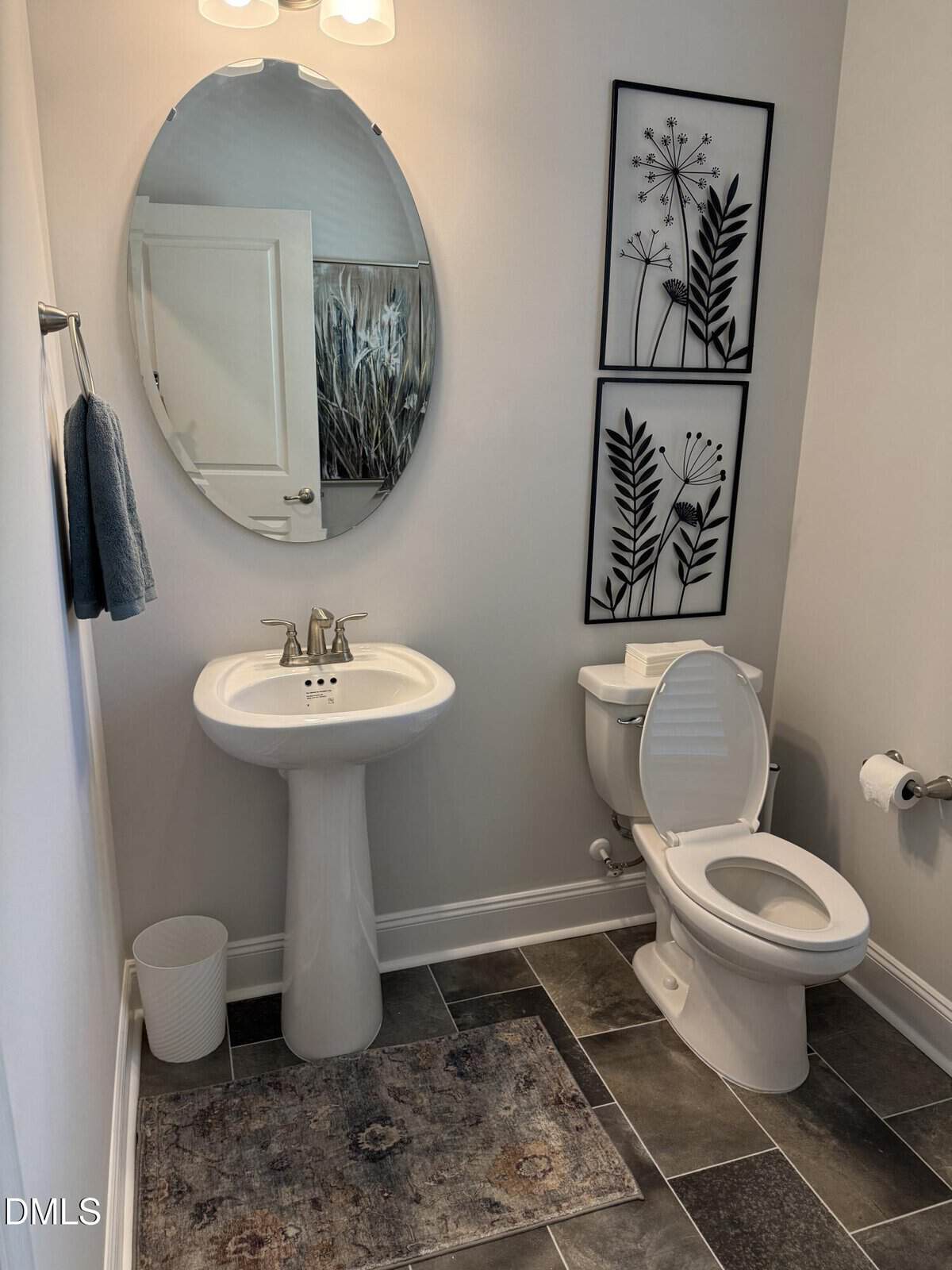 A clean bathroom with a pedestal sink, oval mirror, toilet with the lid open, two black botanical prints on the wall, a wastebasket, a towel on a rack, and a gray patterned rug on a tiled floor.
