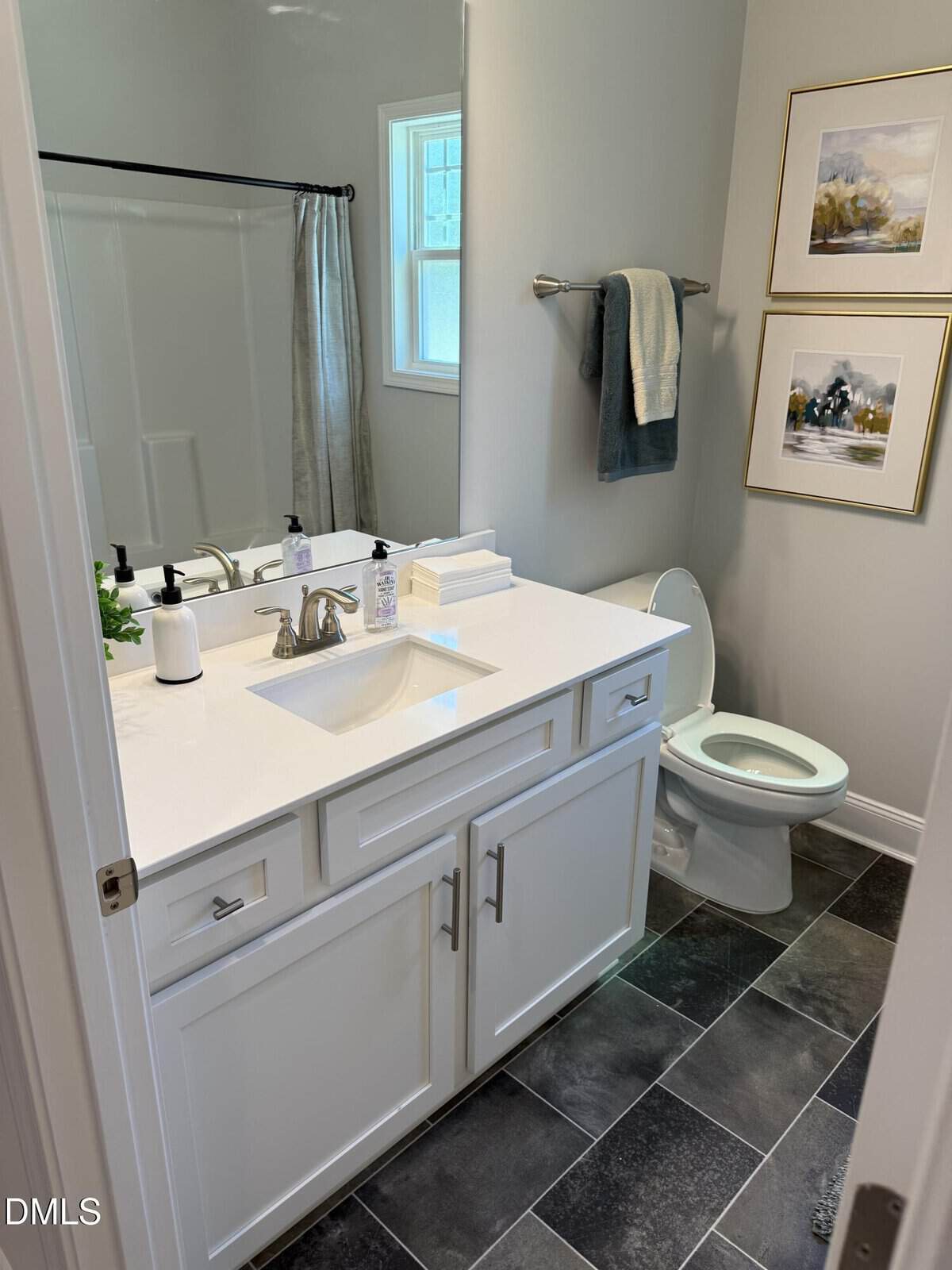 Modern bathroom with white vanity, sink, soap dispenser, towels, toilet, two framed pictures on the wall, window, and dark tile floor. Shower with gray curtain visible in the background.