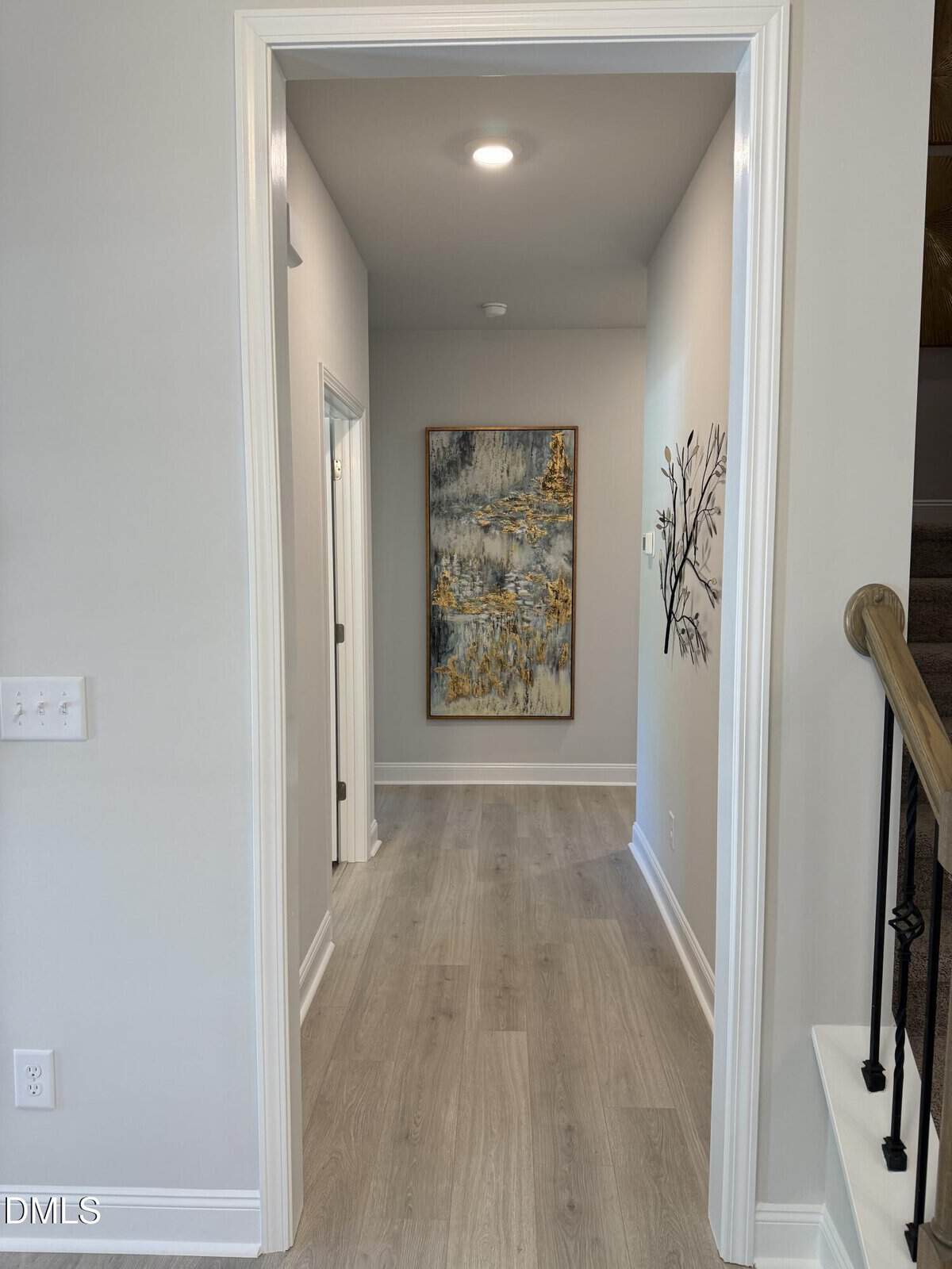 A hallway with light wood flooring, gray walls, and white trim leads to a large abstract painting at the far end. There are two doorways on the left and a staircase with a wooden handrail on the right.