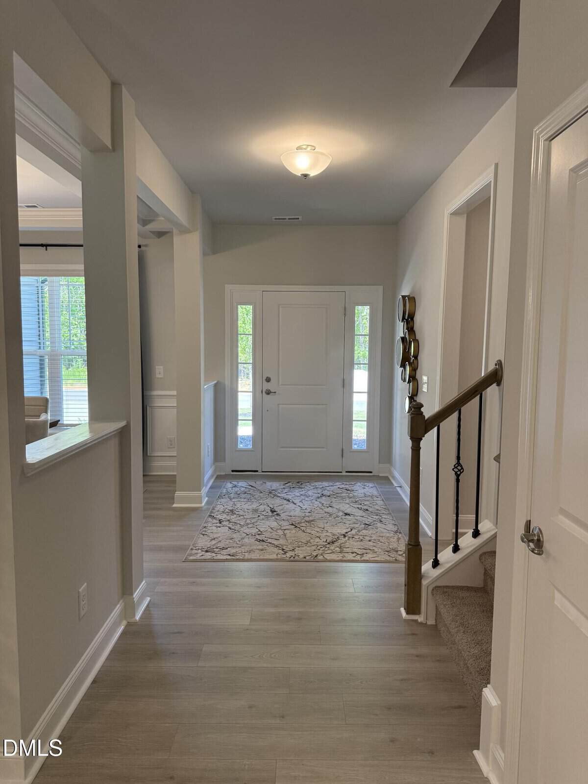 A hallway with light wood flooring leads to a white front door with glass panels. A patterned rug is placed near the entrance, and carpeted stairs with a wooden handrail are on the right. Neutral-colored walls and trim throughout.