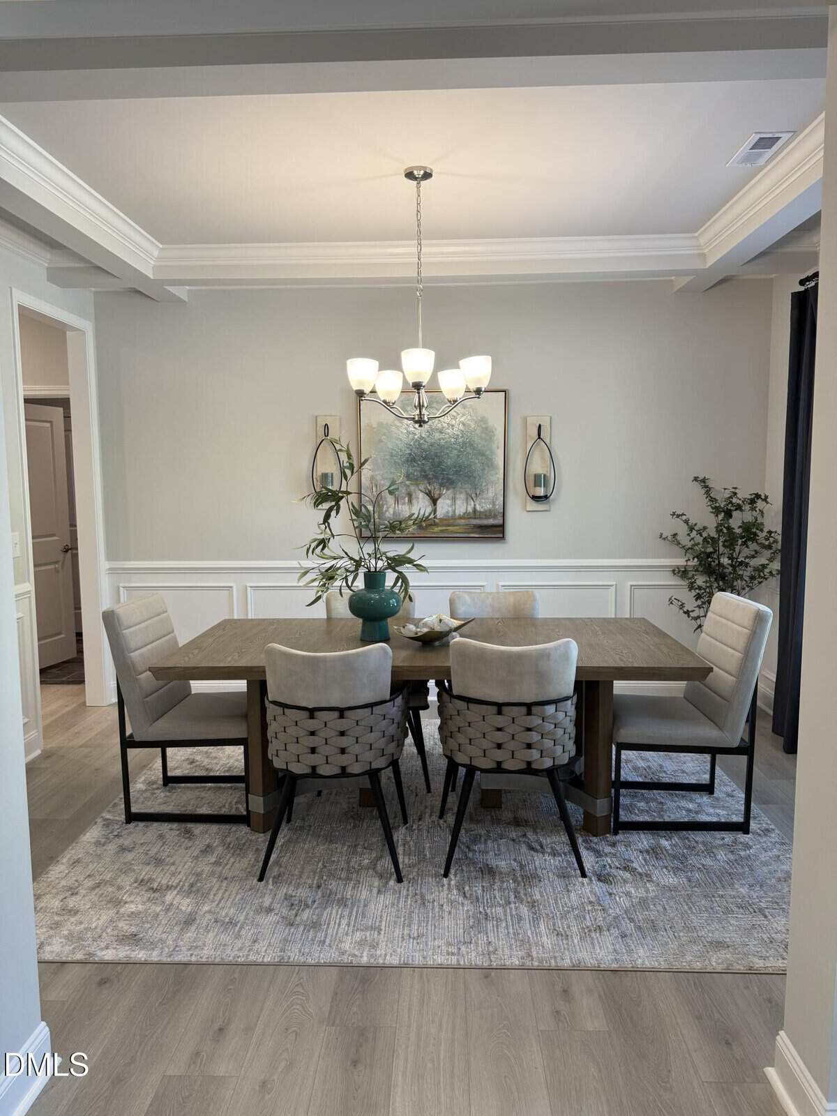 A modern dining room with a wooden table, six upholstered chairs, a large rug, a central teal vase with branches, and a chandelier above. A landscape painting and two wall sconces are on the back wall.