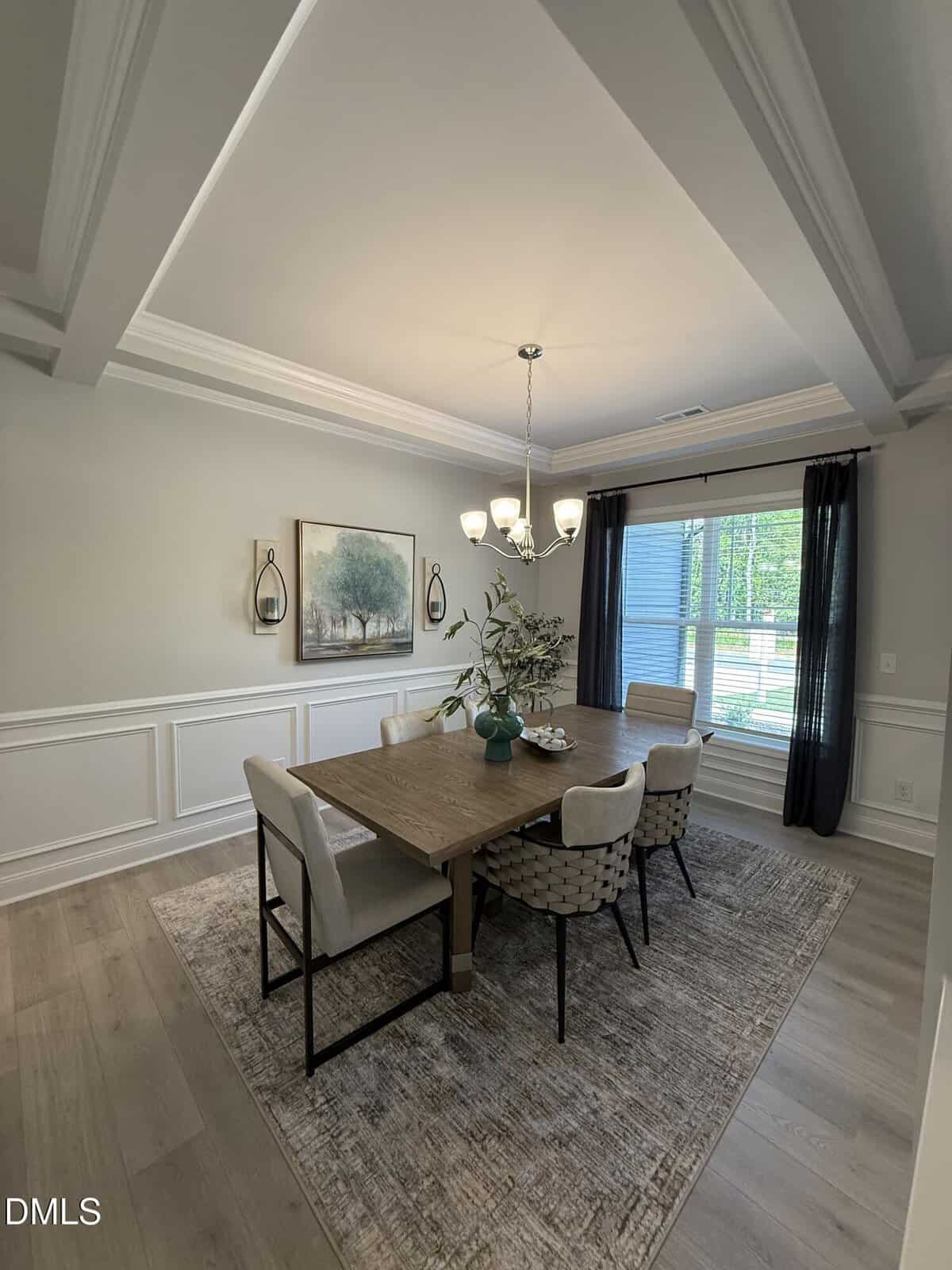 A dining room with a rectangular wooden table, six cushioned chairs, a rug, a modern chandelier, wall art, two sconces, a plant centerpiece, and large windows with dark curtains allowing natural light in.