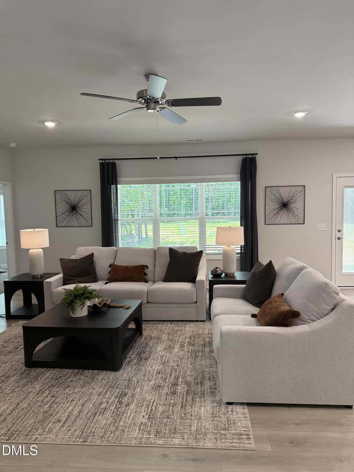 A modern living room with two light gray sofas, a dark wood coffee table, a patterned rug, side tables with lamps, decorative pillows, wall art, and large windows with dark curtains letting in natural light.