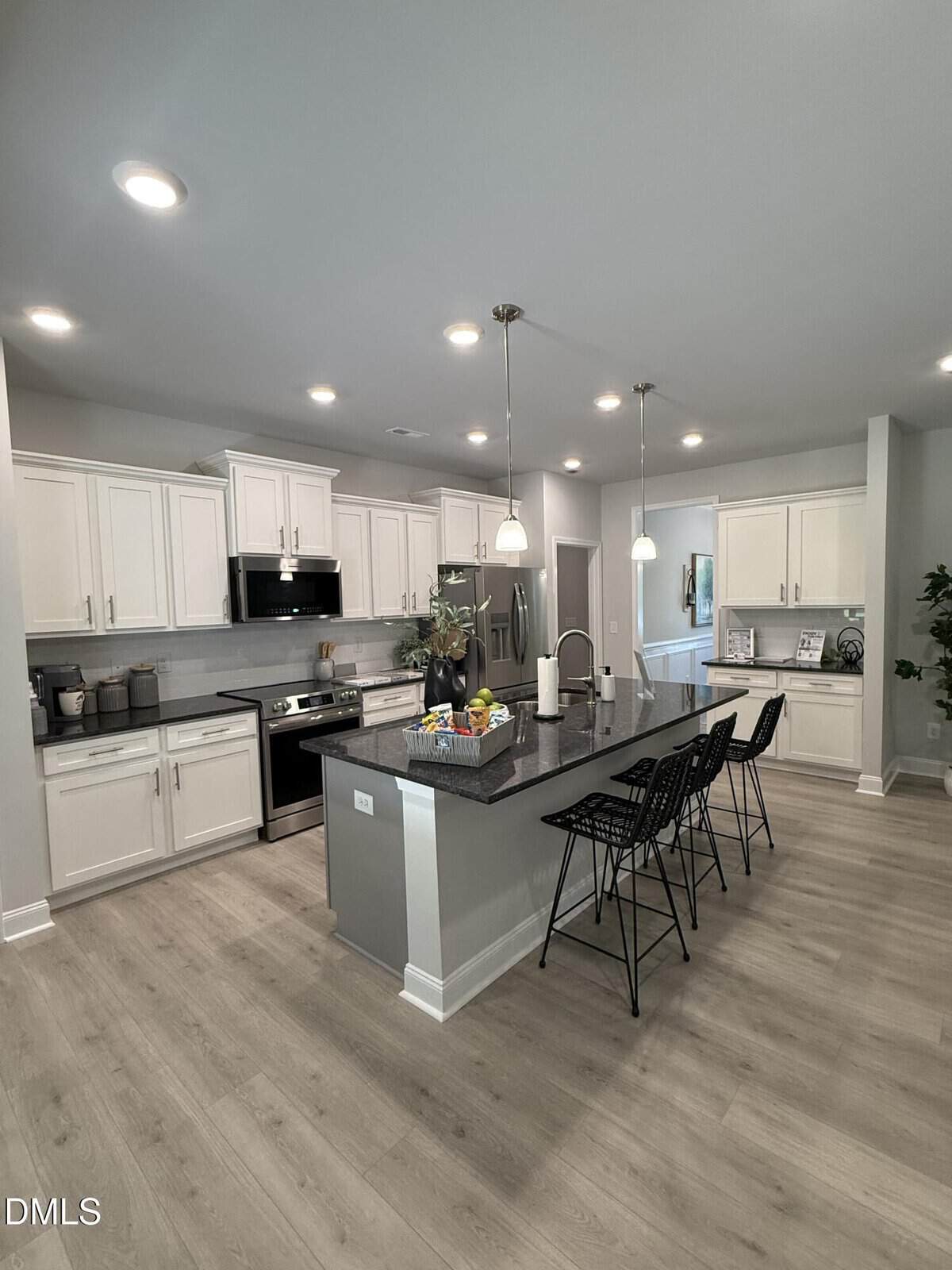 Modern kitchen with white cabinets, stainless steel appliances, a gray island with three black barstools, pendant lighting, and light wood flooring. The island has a fruit bowl and decorative items.