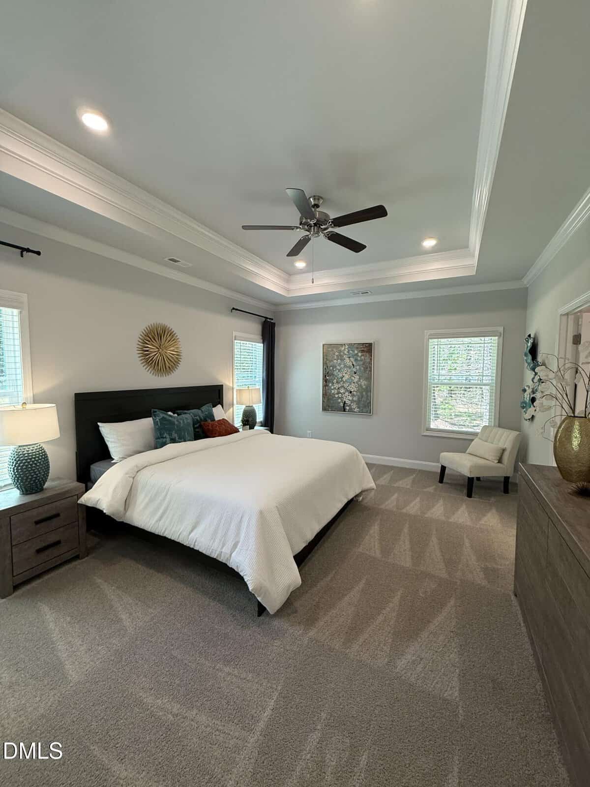 A spacious bedroom with a king-sized bed, white bedding, dark wood furniture, two windows, an accent chair, decorative wall art, a ceiling fan, and neutral-colored carpeting.