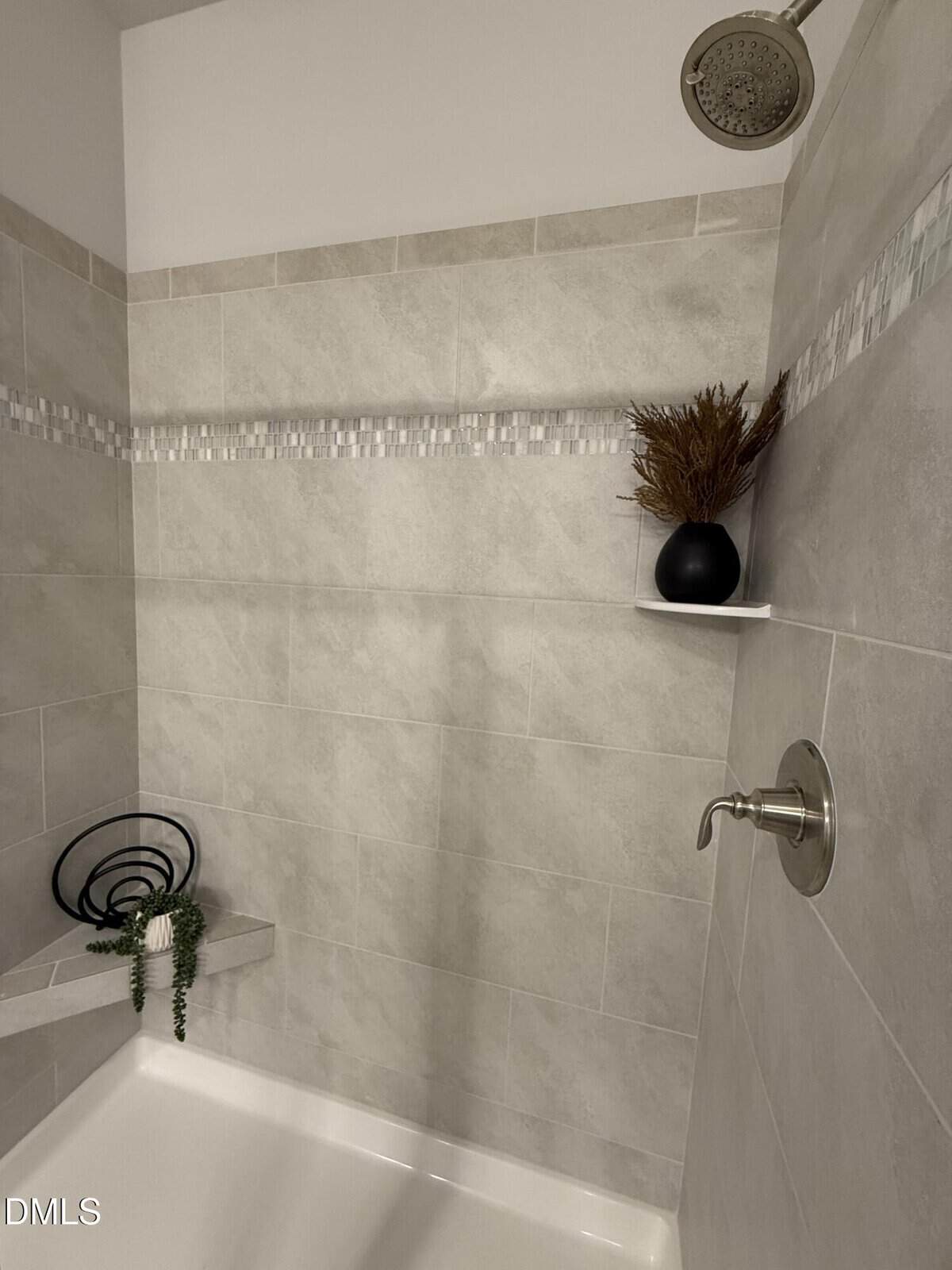 A tiled shower with light gray tiles, a silver showerhead and faucet, a built-in corner seat with a black decorative piece and green plant, and a small shelf holding a black vase with dried foliage.