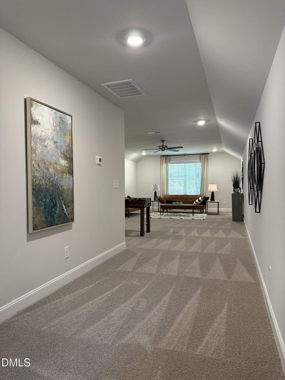 A hallway with carpeted flooring leads to a spacious room featuring a brown sofa, foosball table, modern decor, artwork on the walls, and large window with light blue curtains. The area is well-lit with recessed lighting.