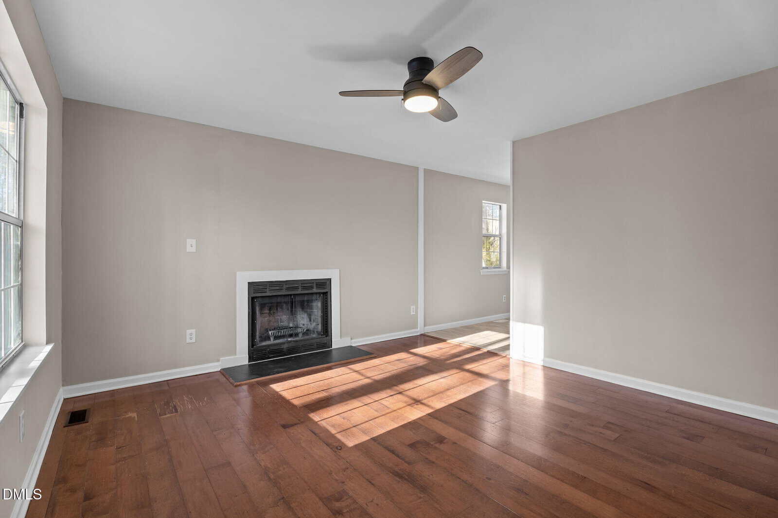 A bright, empty living room with light beige walls, a wood floor, a ceiling fan with a light, a fireplace with a black surround, and large windows letting in natural light.