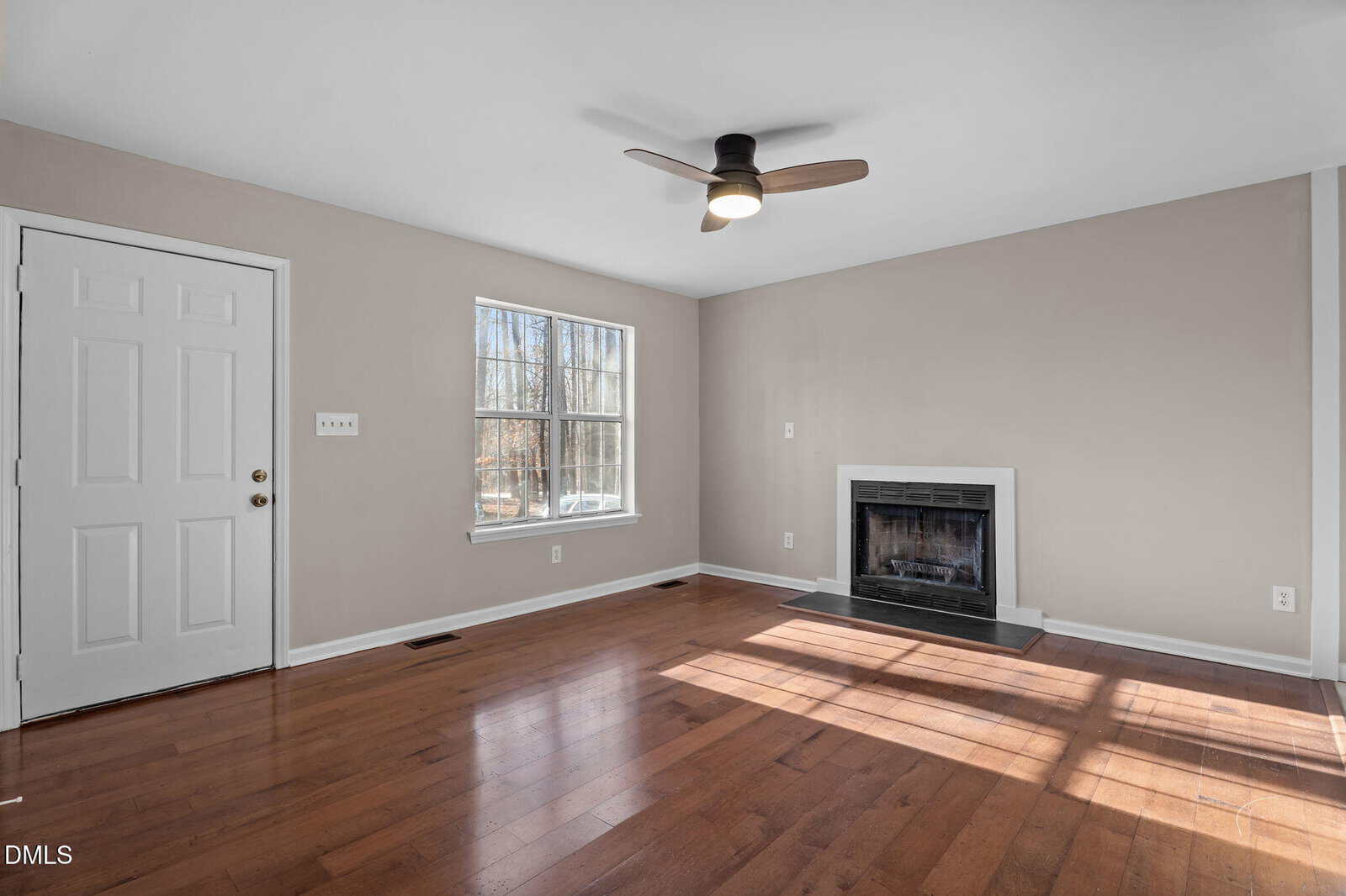 A bright, empty living room with hardwood floors, beige walls, a white door, a window letting in sunlight, a ceiling fan, and a fireplace with a black surround and white mantel.