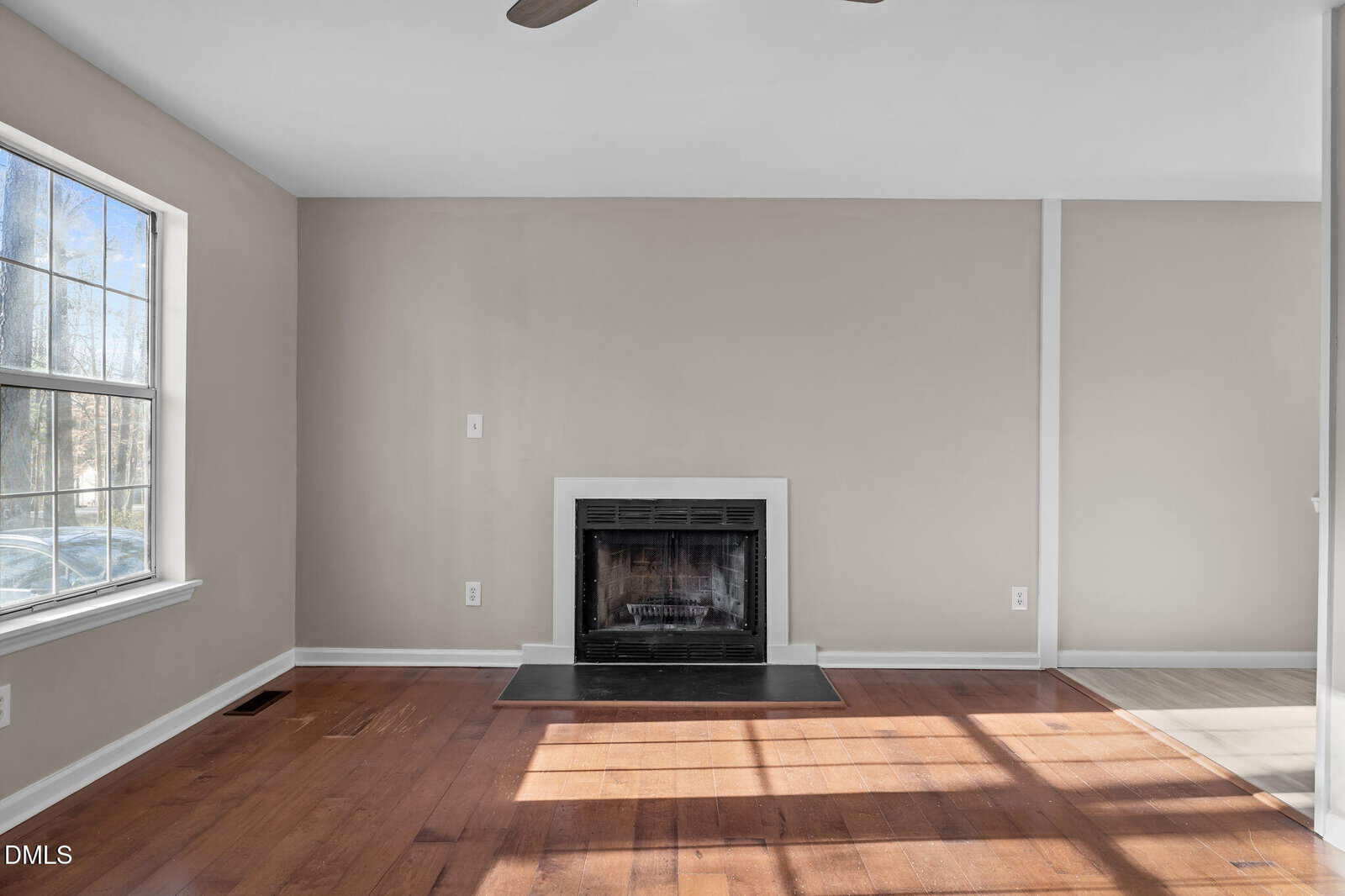 A room with light beige walls, a large window on the left, a black-framed fireplace centered on the back wall, and hardwood floors. Sunlight streams in through the window, casting shadows on the floor.