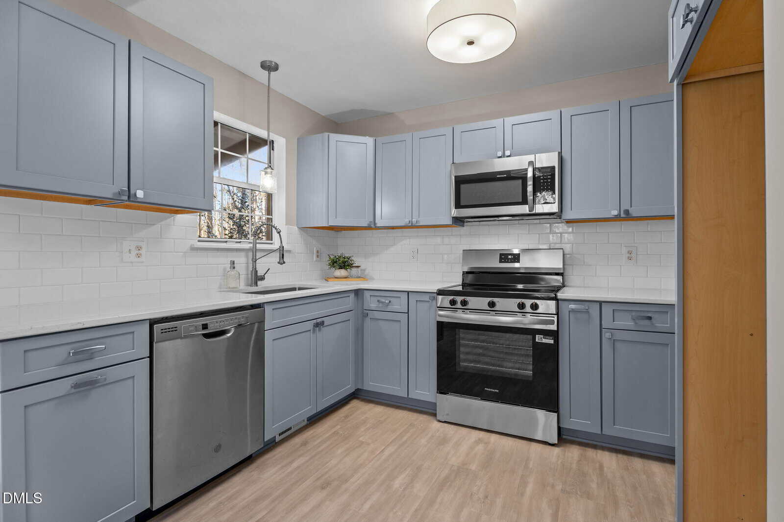Modern kitchen with light blue cabinets, stainless steel appliances including a stove, microwave, and dishwasher, white subway tile backsplash, white countertops, light wood flooring, and a round ceiling light.