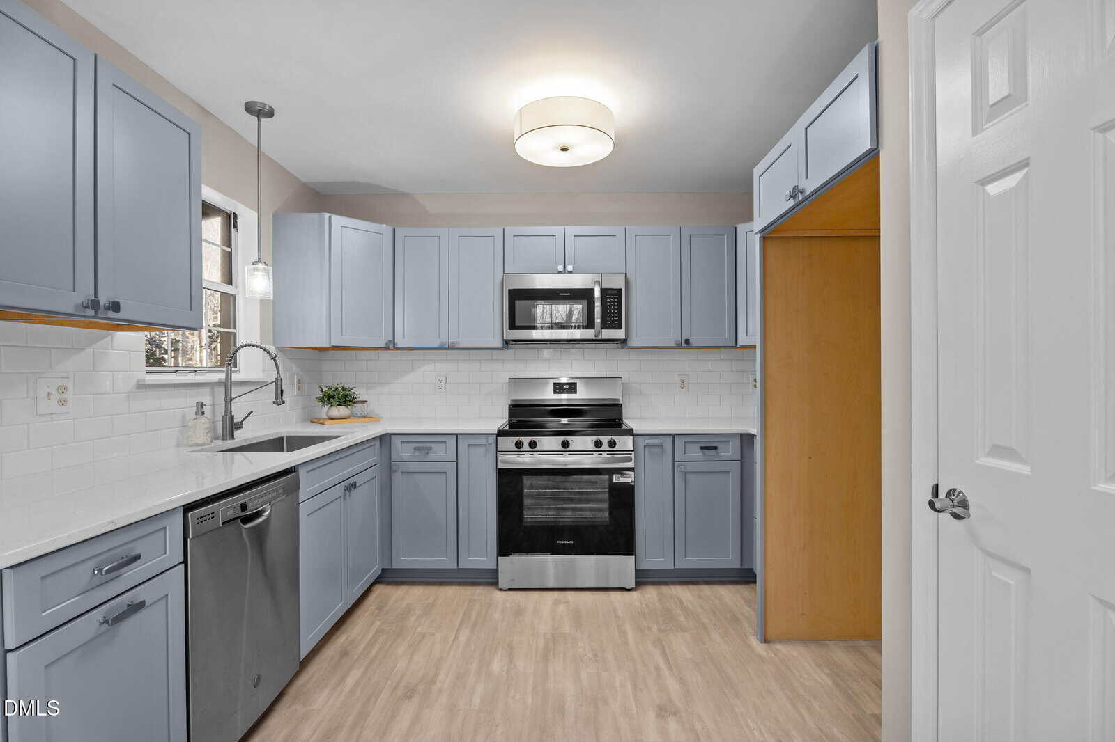 A modern kitchen with light blue cabinets, stainless steel appliances, a white subway tile backsplash, light wood flooring, and a ceiling light fixture. There is an empty pantry space to the right of the stove.