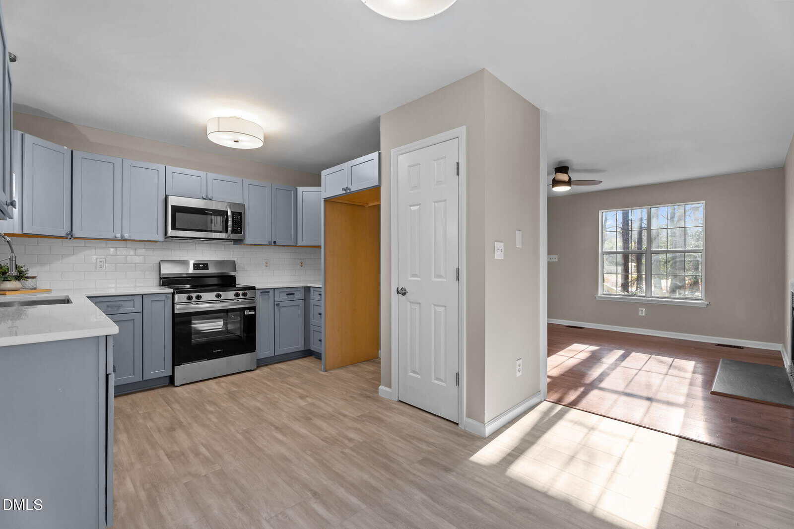 Modern kitchen with light blue cabinets, stainless steel appliances, white subway tile backsplash, and light wood flooring; adjacent room with large window and ceiling fan is visible.