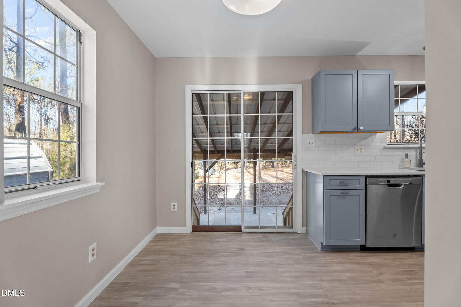 A kitchen with light gray cabinets, a stainless steel dishwasher, white tile backsplash, and wood-style flooring. Large windows and glass sliding doors let in natural light, and a covered patio is visible outside.