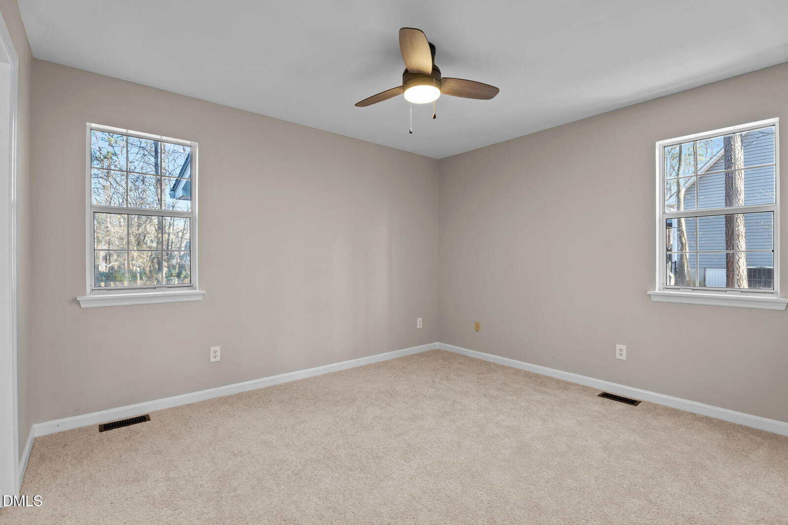 Empty bedroom with beige carpet, light gray walls, two windows, a white baseboard, and a ceiling fan with a light. Daylight shines through the windows, showing trees and a nearby house outside.