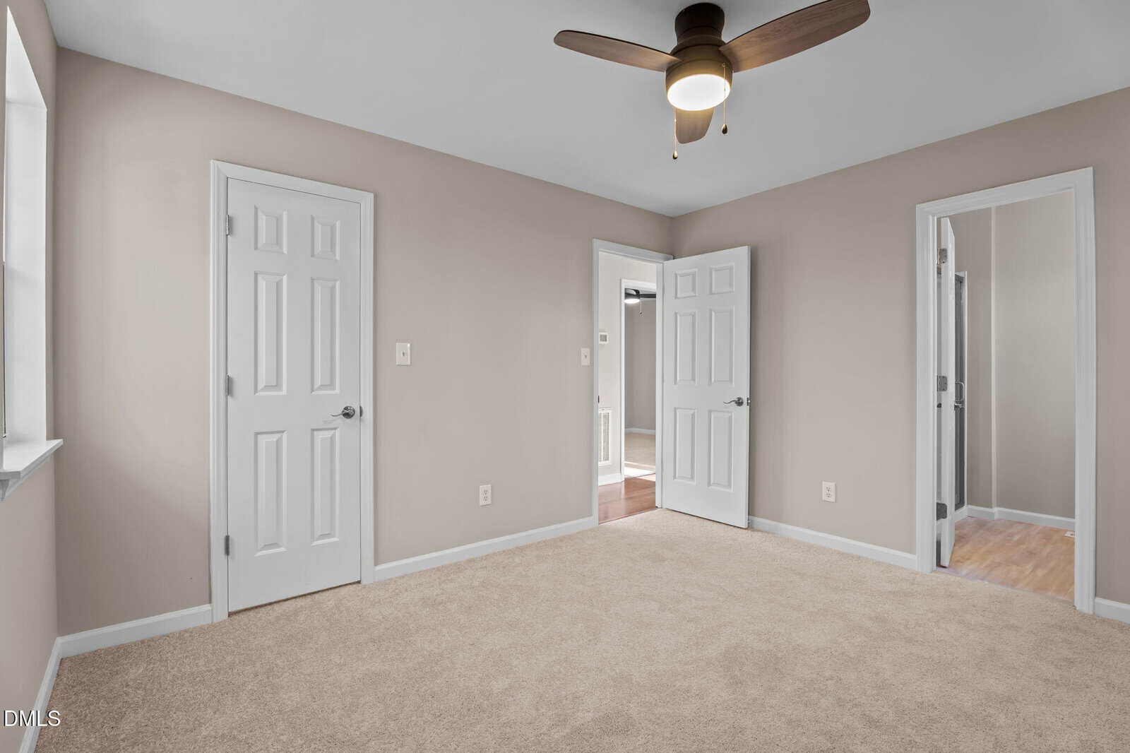 Empty beige carpeted room with tan walls, two closed white doors, one open door, a window, and a ceiling fan with a light.