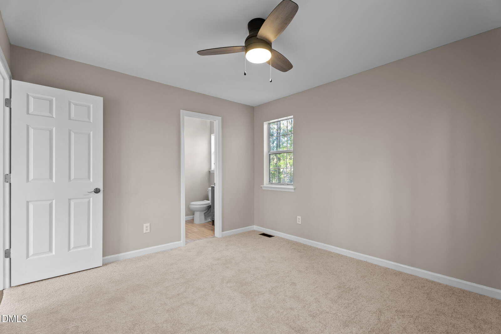 A carpeted bedroom with beige walls, a ceiling fan with lights, a window, an open door, and an open doorway leading to a bathroom with a visible toilet.