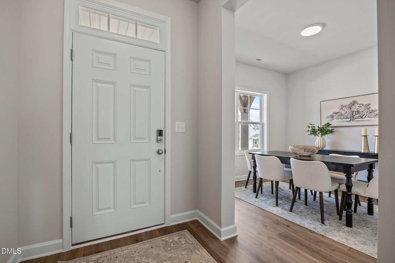 A white front door with a keypad lock opens to a hallway with light-colored walls and wood flooring; to the right, there is a dining room with a dark table, six beige chairs, a rug, and a framed art piece on the wall.