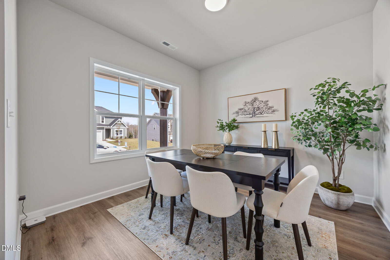 A dining room with a black table, six white chairs, a light patterned rug, large window, sideboard with decor, framed tree artwork, and a tall potted plant in the corner.