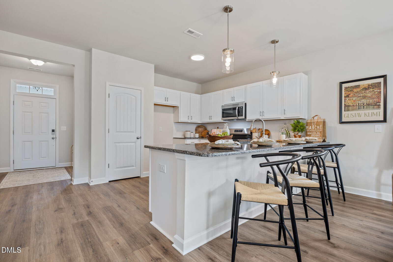 Modern kitchen with white cabinets, stainless steel appliances, and an island with three black chairs. Pendant lights hang above the island. A “Gather Here” sign and framed art decorate the wall. Light wood flooring throughout.