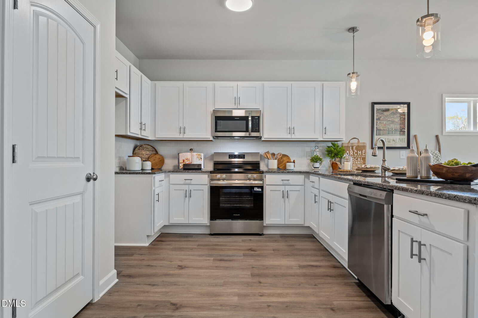 Modern kitchen with white cabinets, stainless steel appliances, granite countertops, and wood flooring. Pendant lights hang above a large island, and various kitchen items and decor are neatly arranged on counters.