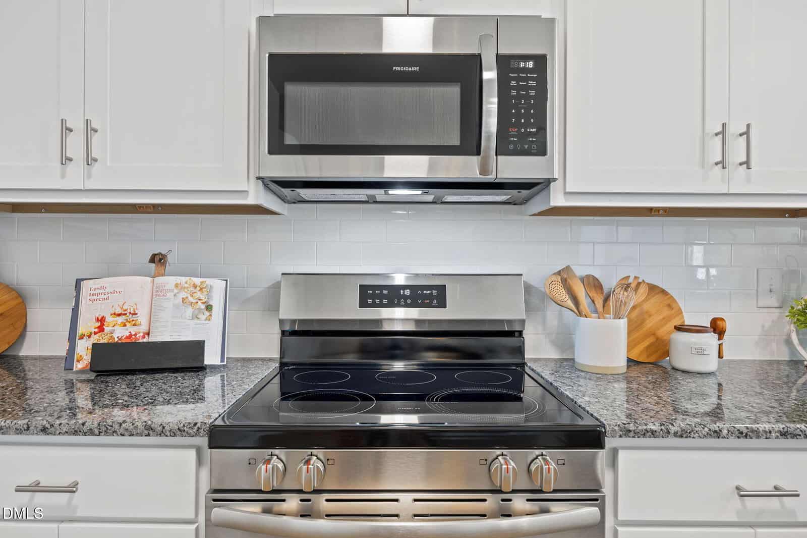 Modern kitchen with a stainless steel stove and microwave, white cabinets, granite countertop, utensils in a white holder, open cookbook, and wooden cutting boards against a white tile backsplash.