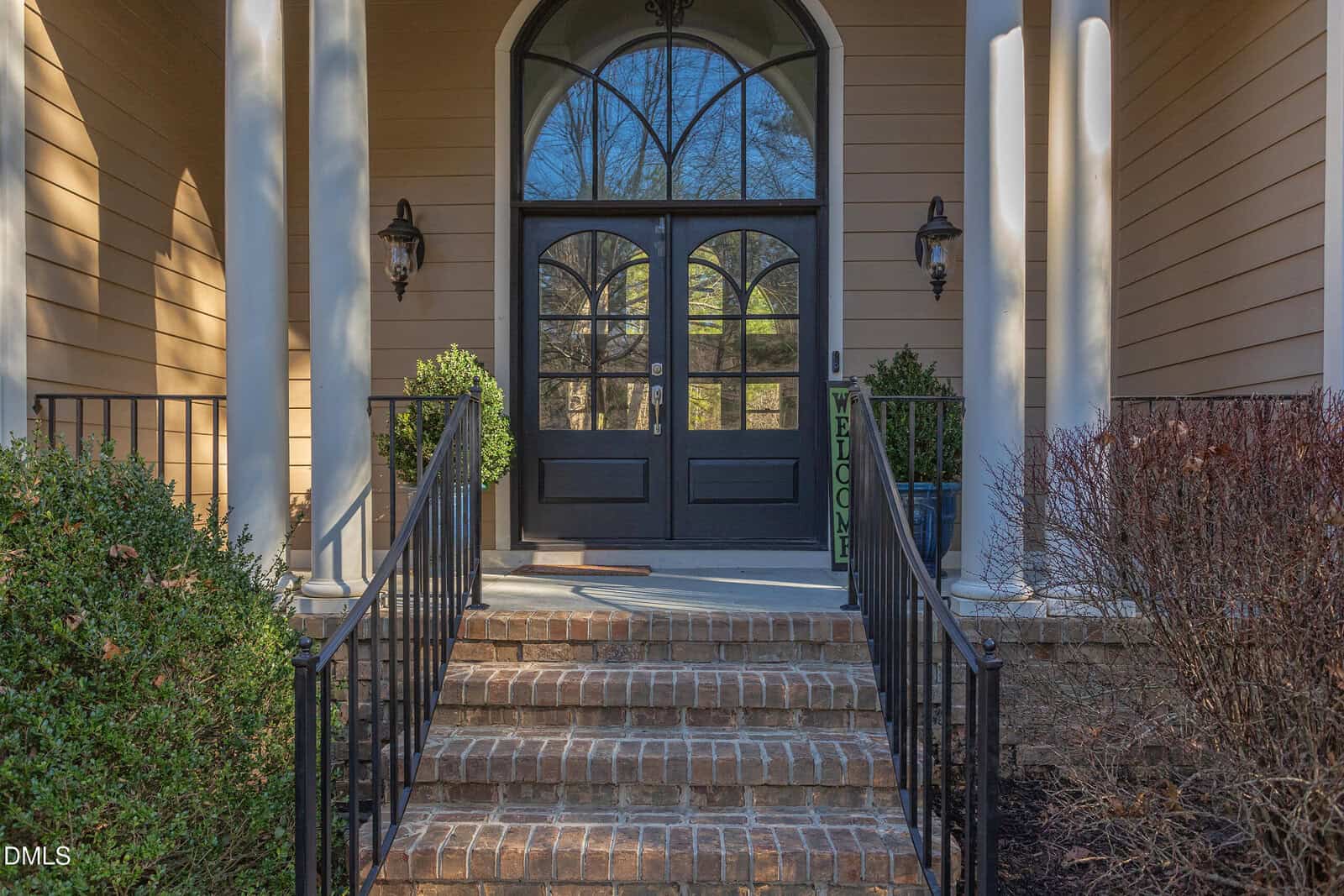 A brick staircase with black railings leads to a double black front door with large arched windows. There are two potted shrubs, exterior wall lanterns, columns, and a vertical "WELCOME" sign by the door.