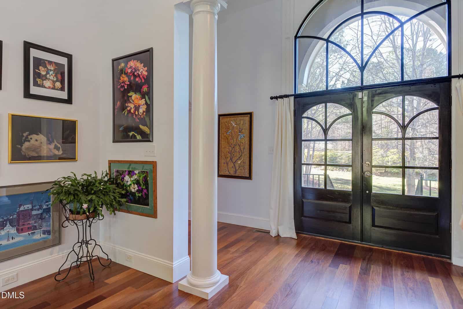 A foyer with dark double doors featuring arched windows, white walls, hardwood floors, a potted plant on a metal stand, and several framed artworks hanging on the wall. Large window shows trees and grass outside.