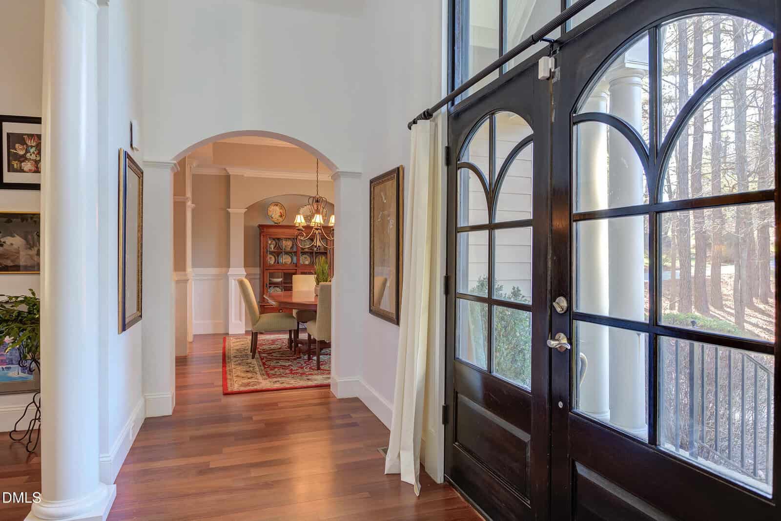 A foyer with dark double doors and white curtains leads to a dining room with a wooden table, chairs, chandelier, and china cabinet. The space features hardwood floors and white trim with large windows letting in natural light.