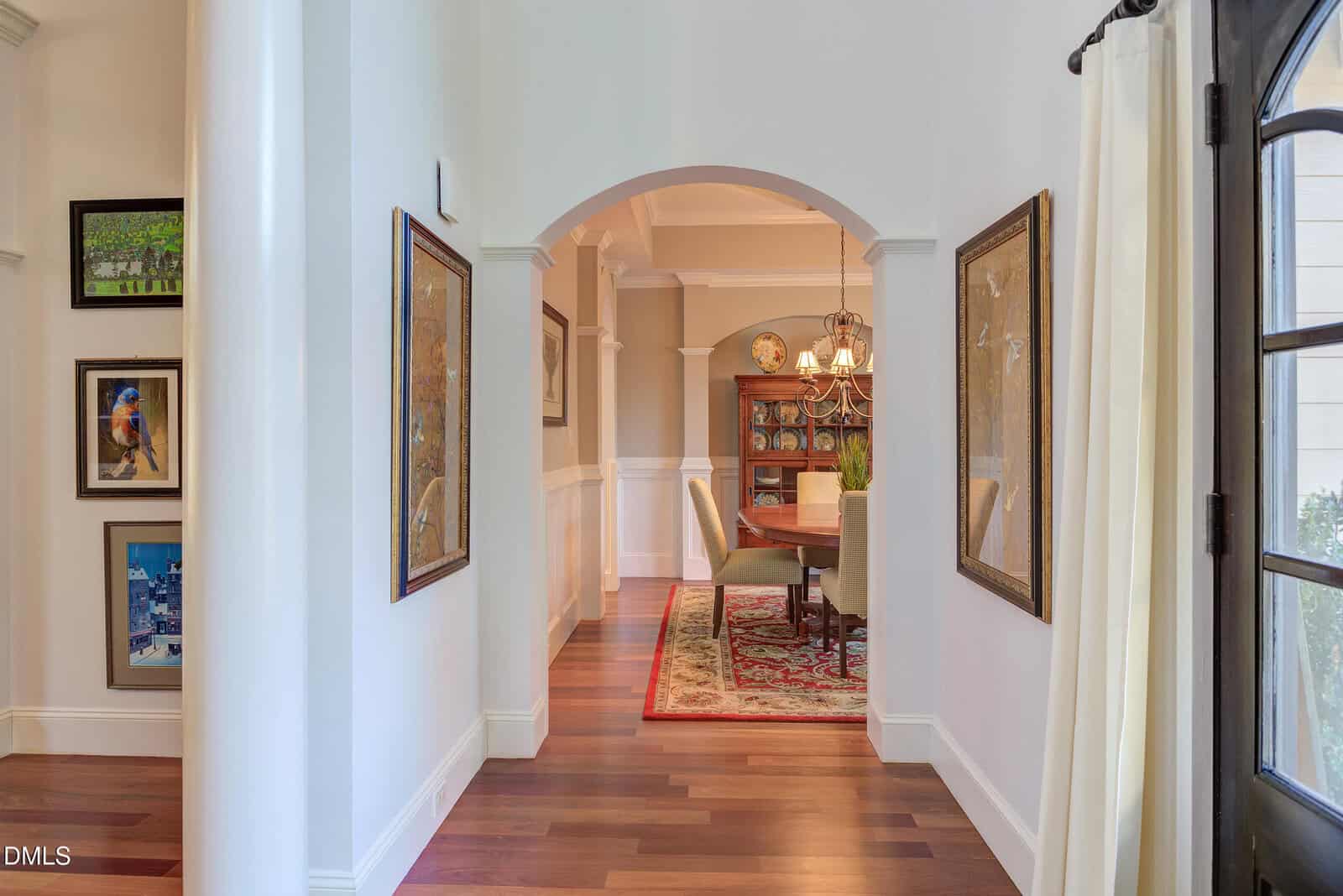 A hallway with framed artwork on white walls leads to a dining room with a wooden floor, a rug, a dining table with six chairs, a chandelier, and a large cabinet against the back wall.