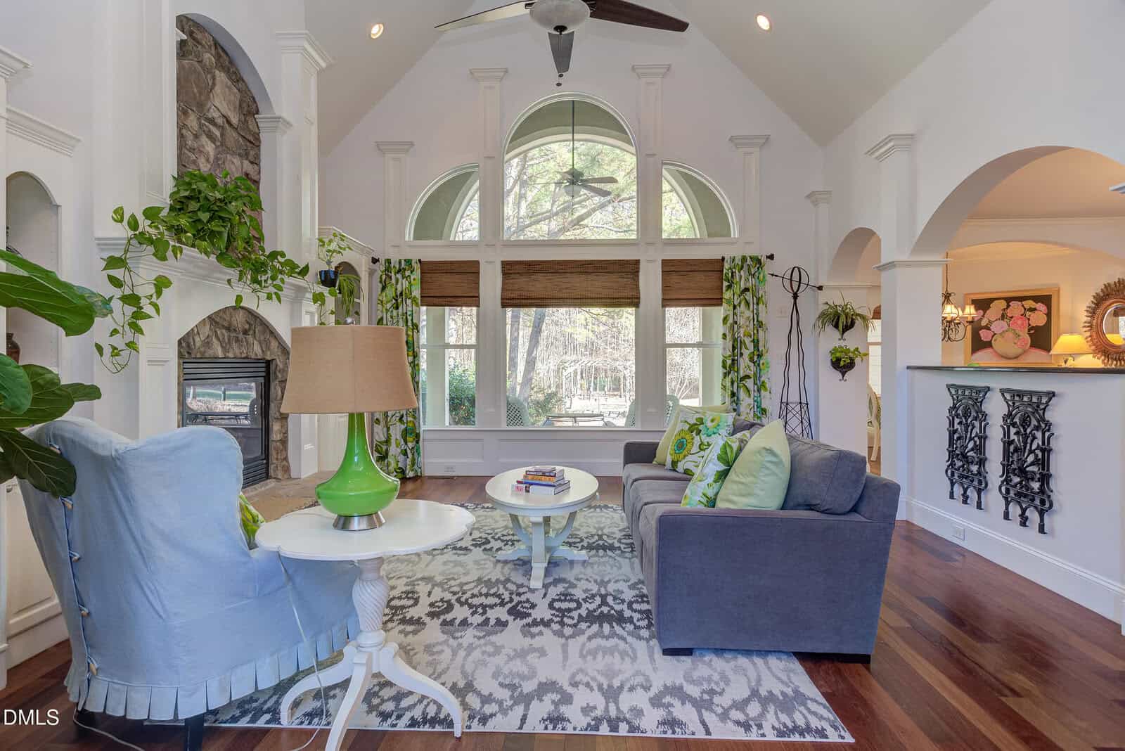 A spacious living room with a high vaulted ceiling, large arched windows, a fireplace, two sofas, a coffee table, and decorative plants. The room features hardwood floors and a patterned area rug.