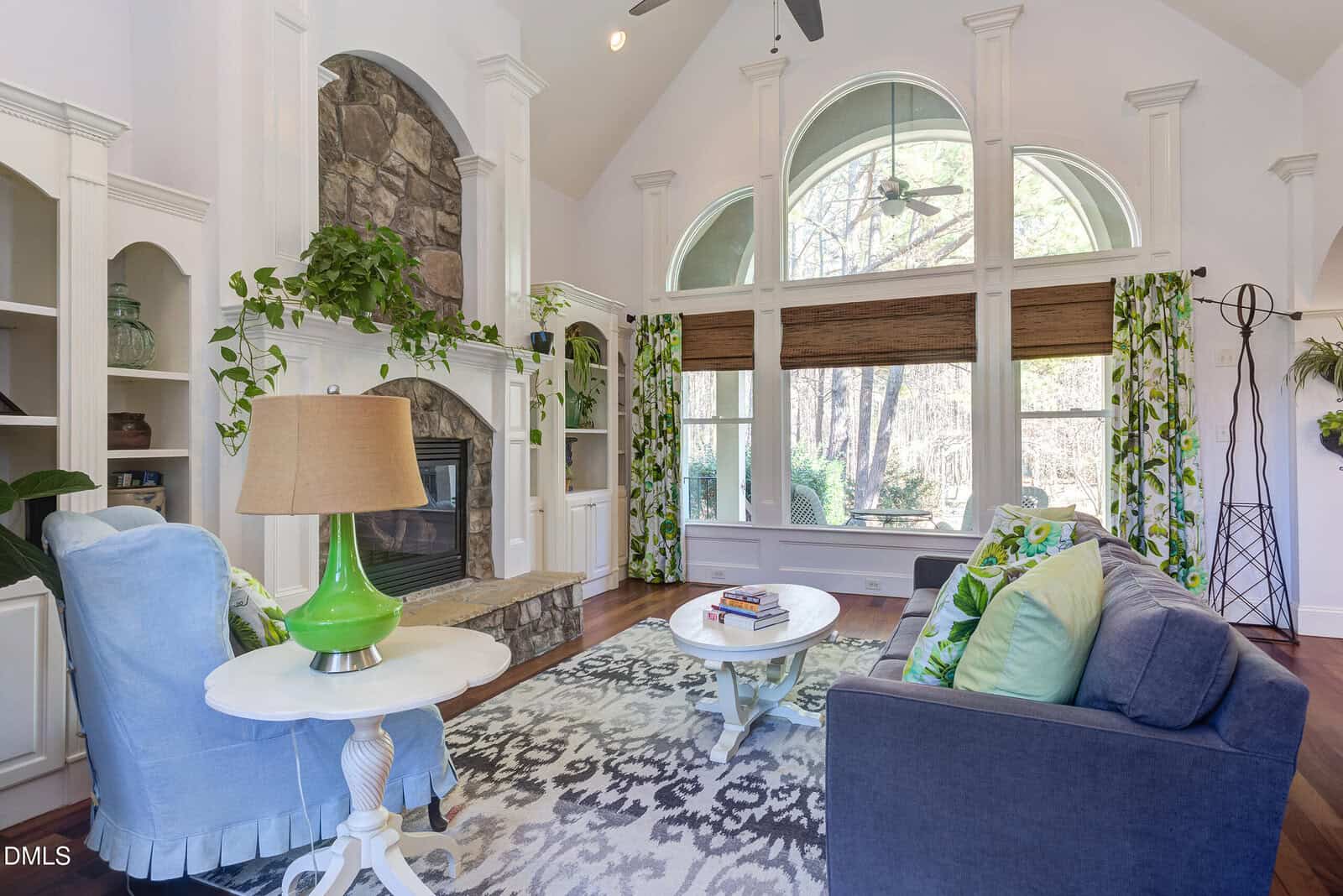 A living room with vaulted ceilings, large arched windows, a stone fireplace, built-in shelves, a gray sofa with pillows, a blue chair, a white coffee table, and green accents including a lamp and curtains.
