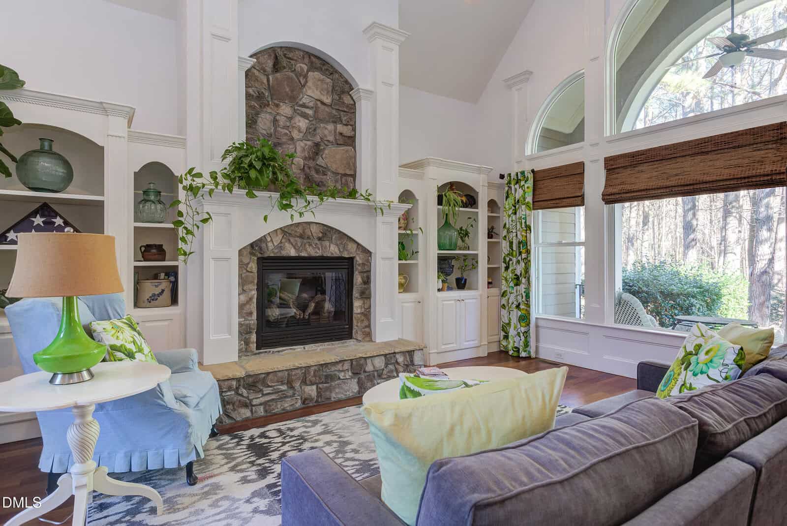 A living room with a stone fireplace, built-in white shelves, a blue armchair, a gray sofa with green and yellow pillows, large arched windows, and hardwood floors. Plants and decorative items are on the shelves.